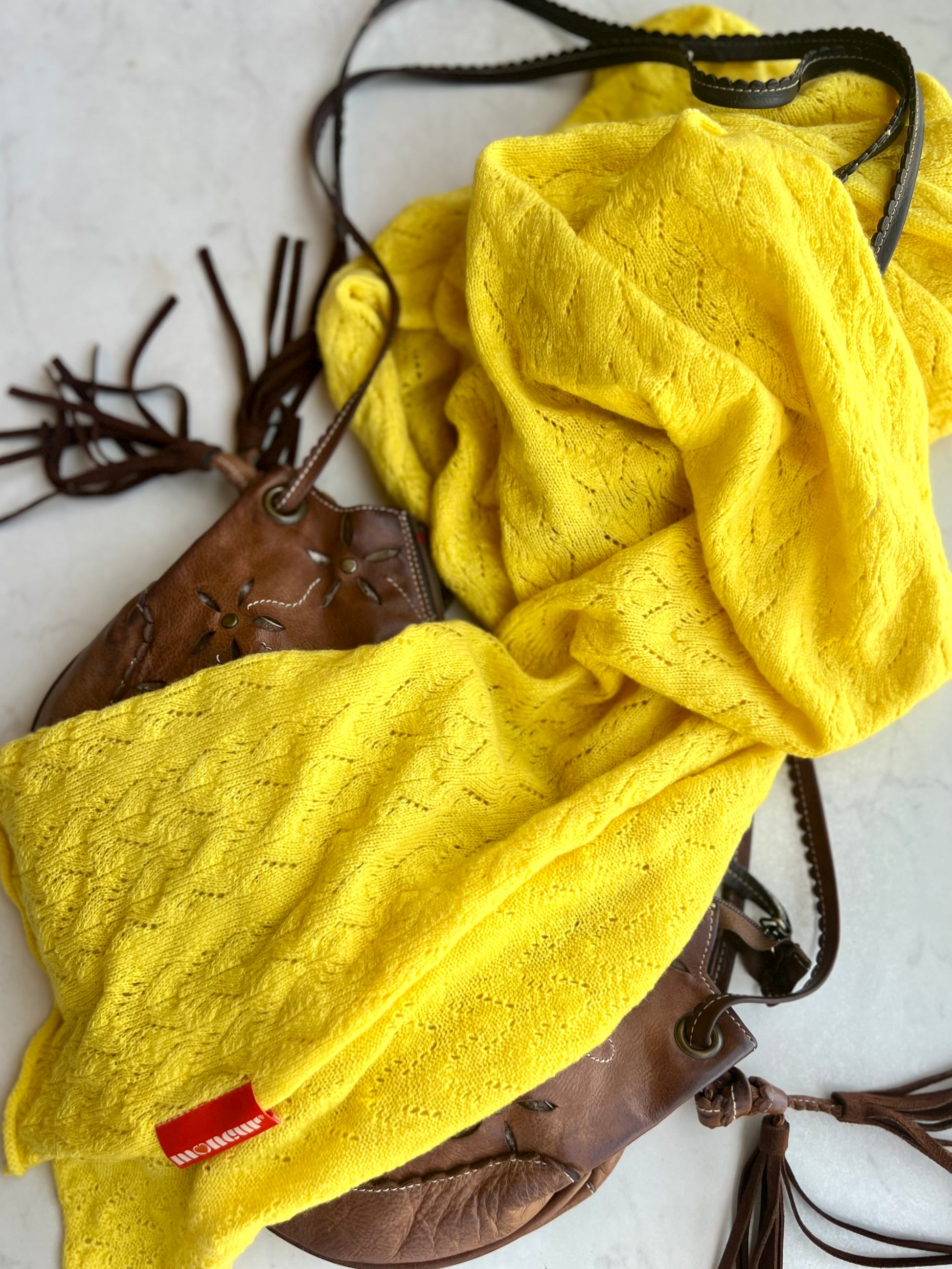 A bright yellow cashmere lace knit wrap laid out on a surface with a soft brown leather bag as if on a sunny holiday.